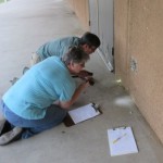 Man and woman kneeling on the ground beside a wall inspecting the base of the wall with flashlights 